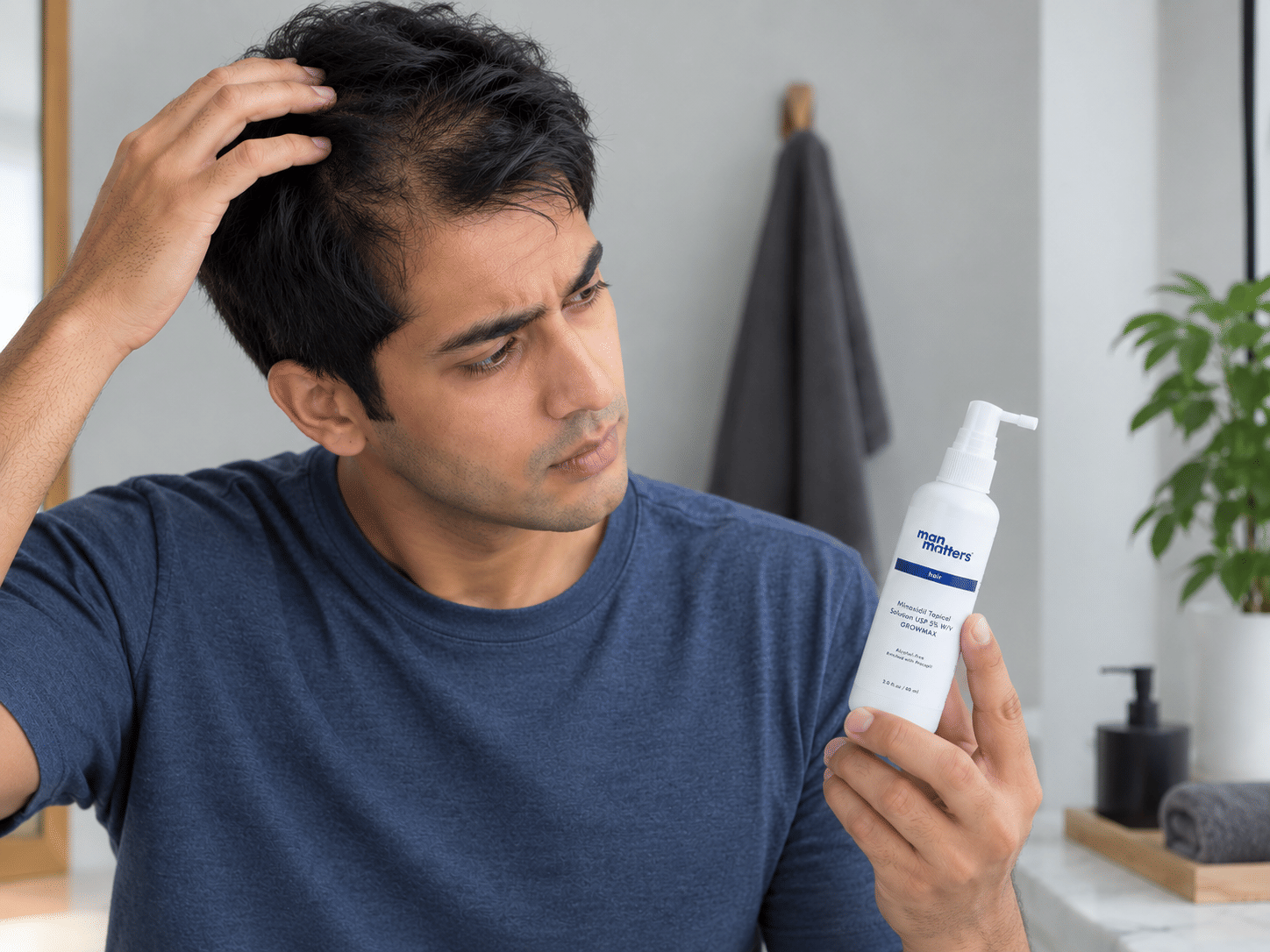 Young Indian man examining his thinning hair while holding a Minoxidil bottle, concerned about hair shedding.
